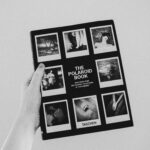 Monochrome image of a hand holding a Polaroid photo book featuring diverse photographs.